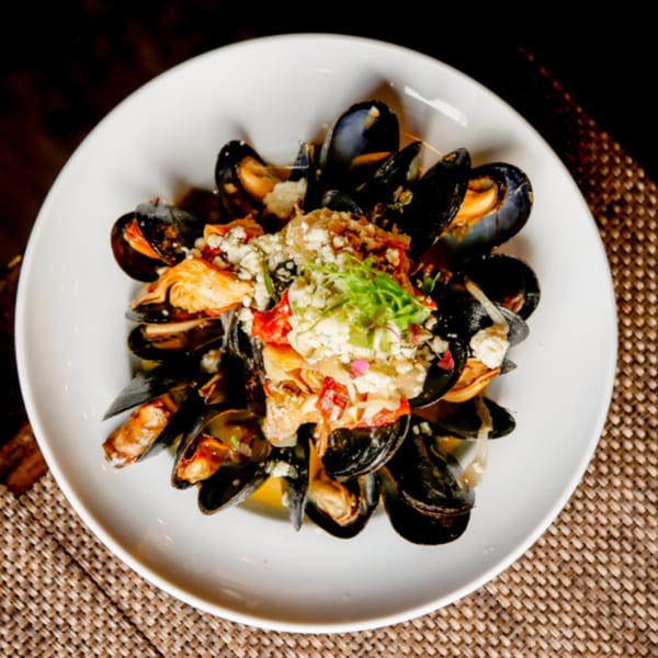 A bistro dish of mussels in a white bowl, topped with crumbled cheese, red peppers, and greens.