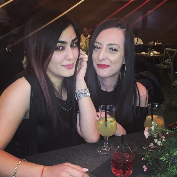 Two women with dark hair and bright lipstick pose with cocktails at a dimly lit bistro.