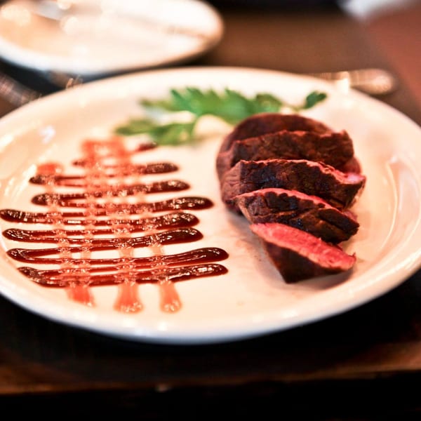 Sliced steak with a decorative sauce drizzle and a sprig of parsley on a white plate.