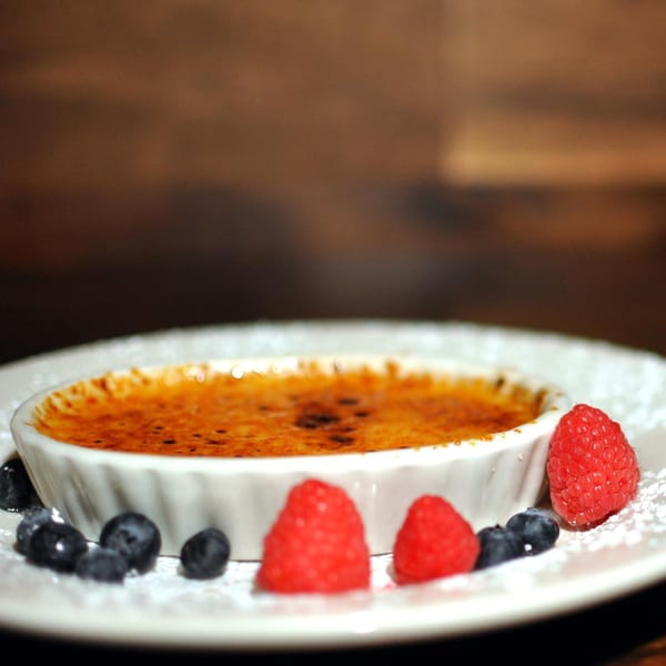 A creme brulee in a white ramekin, garnished with raspberries, blueberries, and powdered sugar.