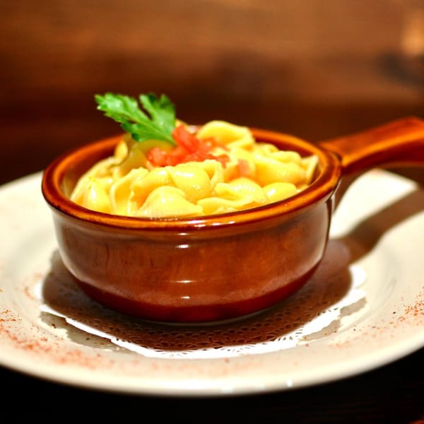 A small brown ceramic pot filled with aged. bistro mac and cheese, garnished with parsley and diced tomatoes.