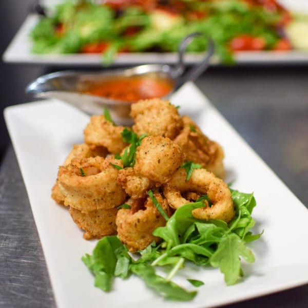 Crispy fried calamari with fresh greens and a side of red dipping sauce on a white plate.