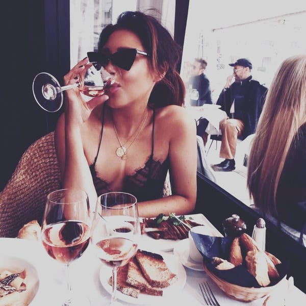 A woman in sunglasses sips rosé wine at a bistro table with food and other diners.