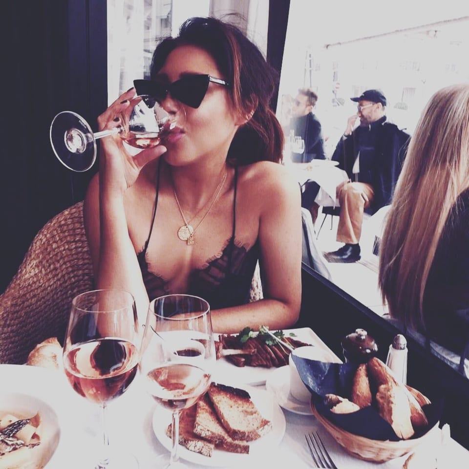 A woman in sunglasses sips rosé wine at a bistro table with food and other diners.