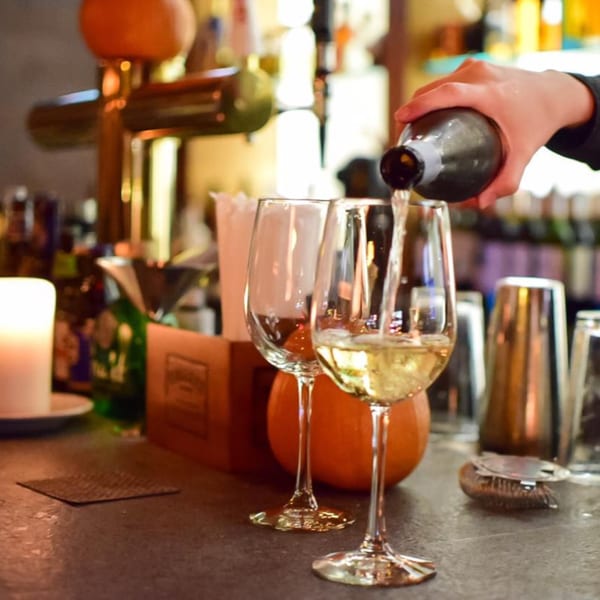 A bartender pours white wine into a glass at a bistro bar, with a candle and pumpkins nearby.