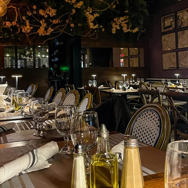 Elegant bistro dining room with set tables, woven chairs, and decorative floral branches overhead.