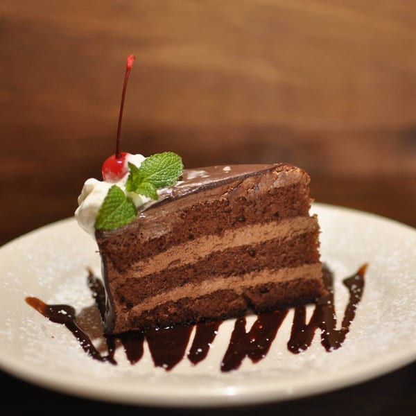 A slice of rich chocolate cake with whipped cream, a cherry, and mint, drizzled with chocolate sauce.
