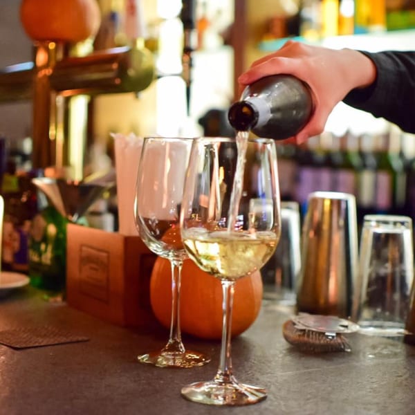 A bartender pours white wine into a glass on a bistro bar, with another empty glass nearby.
