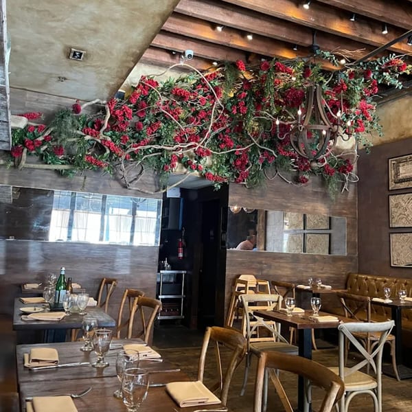 Aged bistro interior with wooden tables, chairs, and a ceiling adorned with red flowers and greenery.