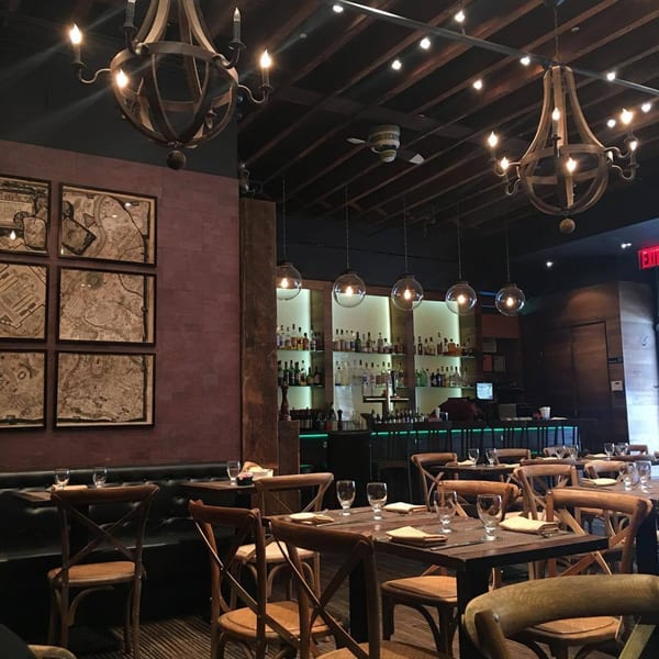 Aged bistro interior with wooden chandeliers, framed maps, and a bar with globe pendant lights.