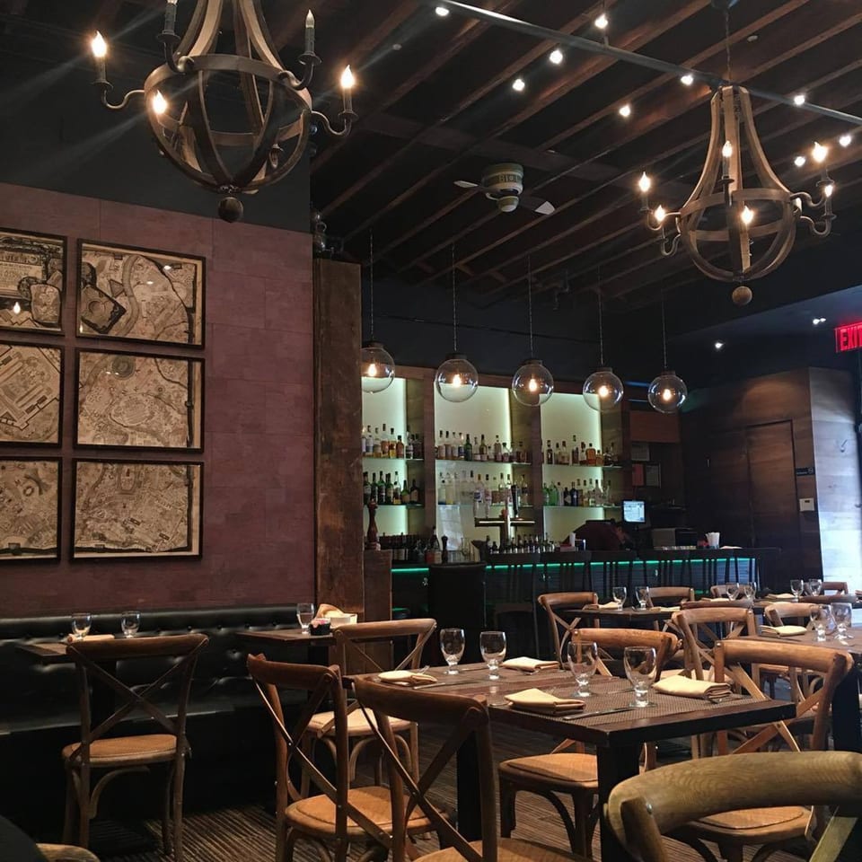 Aged bistro interior with wooden chandeliers, framed maps, and a bar with globe pendant lights.